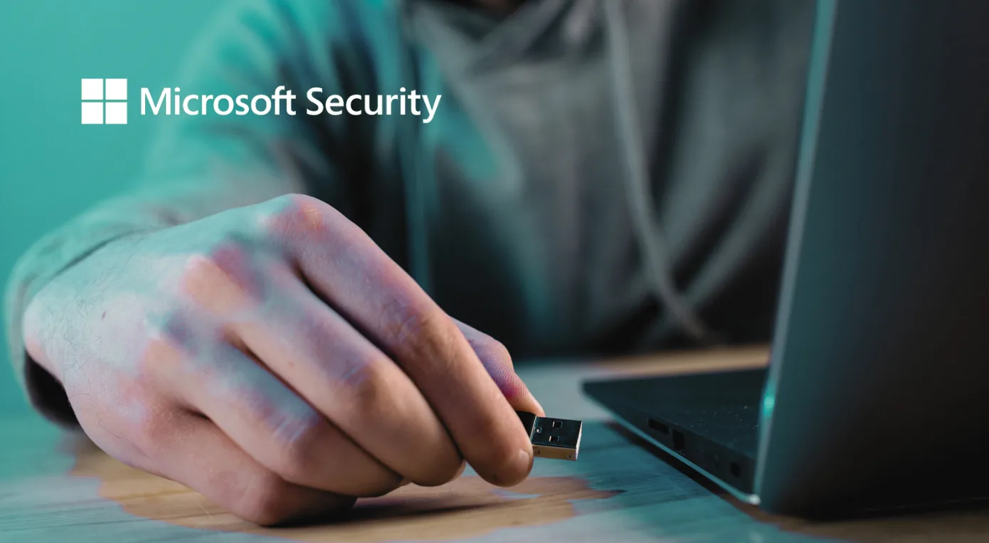 Mão segurando um pen drive USB diante de um notebook, em imagem com a marca Microsoft Security