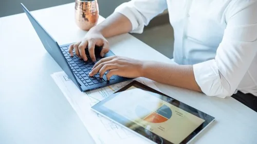 Pessoa digitando em um notebook, com um tablet ao lado exibindo um gráfico de pizza